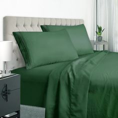 a bed with green sheets and pillows next to a night stand on a nightstand in front of a window