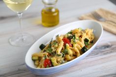 a pasta dish with tomatoes, zucchini and spinach in a white bowl next to a glass of wine