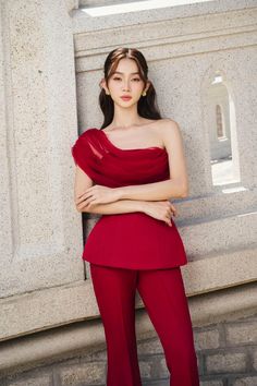 a woman standing in front of a stone wall wearing a red one shoulder top and matching pants