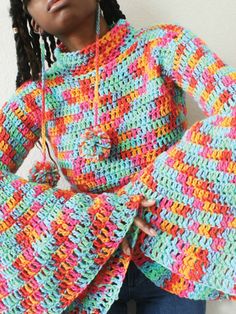 a woman wearing a multicolored crocheted sweater and earphones standing in front of a white wall