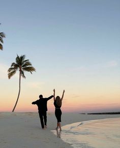 a man and woman walking on the beach with palm trees in the background at sunset