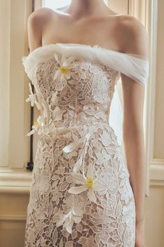 a woman in a white dress with flowers on it's chest and shoulder, standing next to a window