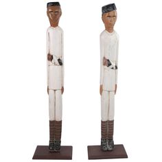 two wooden statues of men in white clothing with black hair and brown boots, one holding a cat