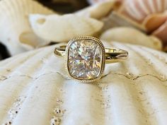 a cushion cut diamond ring sitting on top of a white sea urn with seashells in the background