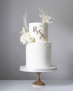 a white wedding cake with flowers and initials on top is sitting on a stand in front of a gray wall