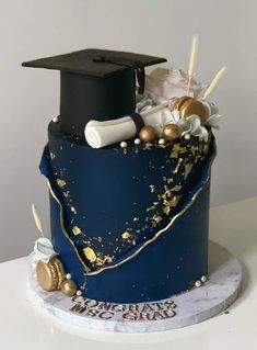 a blue graduation cake with gold and white decorations on it's side, topped with a black cap