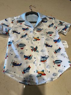 Classy crisp Airplane-Themed button-down shirt for either boys or girls.  What could be better than a nice, button-down shirt?  This version is made up of 100% white cotton printed with airplanes, clouds, helicopters, and stars  The inside yoke is made of 100% cotton blue and white striped fabric.  The shirt-tails are rounded for a more relaxed look. Available in a child's size 8 and Baby size 18-24 months.  Matching striped shorts available in size 18-24 elsewhere in this shop. Collared Shirt For School In Summer, Collared Summer Shirt For School, Cute White Button-up Shirt, Playful Collared Shirt For Summer, Cute White Button Shirt, Short Sleeve Shirt For Playtime, White Printed Button-up Short Sleeve Shirt, White Collared Shirt With Snap Buttons, Cute Short Sleeve Shirt With Buttons