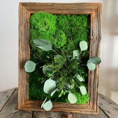 a wooden frame with moss and leaves in it on top of a table next to a plant