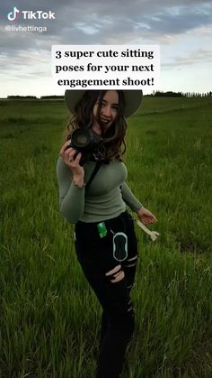 a woman is standing in the grass holding a camera and pointing at her right hand