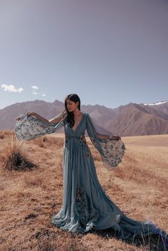 a woman in a long dress standing on top of a dry grass covered field with mountains in the background