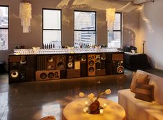 a living room filled with lots of furniture and speakers on top of a table in front of two large windows