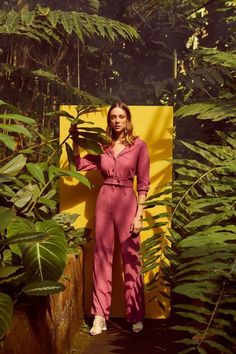 a woman in pink jumpsuits standing next to green plants and yellow wall with trees