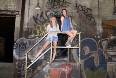 two young women sitting on top of a metal hand rail in front of graffiti covered wall