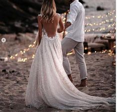 a man and woman standing on the beach holding hands with lights in the water behind them