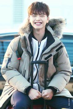 a young man sitting on top of a piece of luggage wearing a coat and smiling