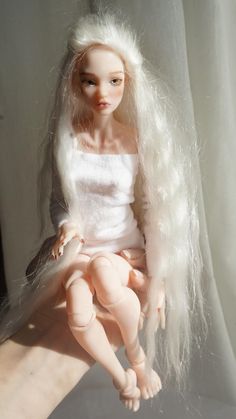 a doll with white hair sitting on top of a persons hand next to a curtain