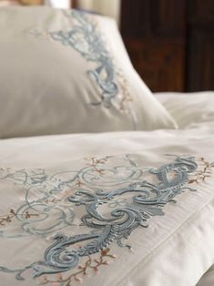 a close up of a bed with white sheets and blue embroidered designs on the pillows