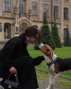 a woman kneeling down next to a dog and kissing her on the nose, with a large building in the background