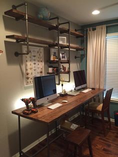 a desk with two computers on top of it in front of a window and some shelves