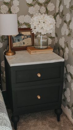 a nightstand with flowers on it next to a wallpapered wall and a lamp