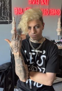 a man with blonde hair and tattoos sitting in front of a neon sign holding his hand up