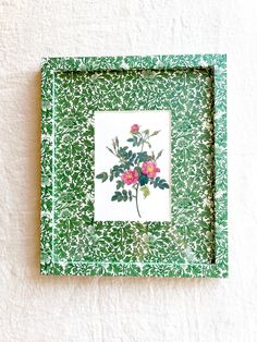 a green frame with pink flowers in it on a white wall next to a black cat