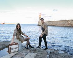 a man and woman sitting on rocks next to the water with a suitcase in front of them