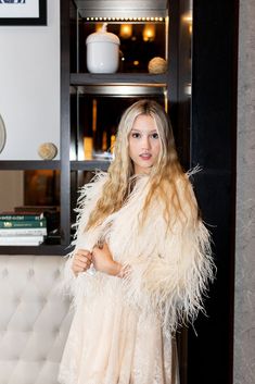 a woman standing in front of a bookshelf wearing a white dress and fur coat