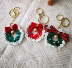 three christmas wreath keychains with bells on them sitting on a white blanket next to a teddy bear