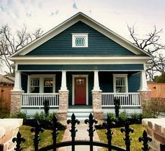 a small blue and white house with a red door on the front porch, surrounded by black iron fence