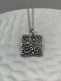 This minimalist tiny rose necklace has been entirely handmade using .999 fine silver. It has been cut out from fine silver precious metal clay, textured, fired and oxidized. The pendant hangs from a sterling silver diamond cut cable chain. This fine silver pendant features an embossed floral rose textured pattern on one side of the pendant and is reversible to a completely different embossed rose pattern on the reverse side. Pendant size: approximately 1/2 x 1/2 inches Chain length: adjustable a Unique Etched Sterling Silver Necklace, Unique Silver Etched Necklaces, Etched Sterling Silver Necklaces, Silver Etched Sterling Silver Necklace, Silver Etched Necklace With Flower Pendant, Artisan Sterling Silver Square Pendant Jewelry, Handmade Square Pendant Jewelry For Everyday, Etched Sterling Silver Pendant Necklace, Artisan Silver Etched Necklace