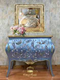 a blue dresser with flowers on it in front of a mirror and a gold frame