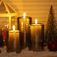 three lit candles sitting on top of snow covered ground next to christmas trees and ornaments