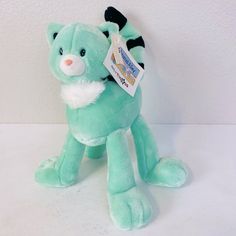 a green teddy bear with a tag on it's chest sitting against a white wall