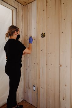 a woman in black shirt and blue gloves working on wood paneled wall with window