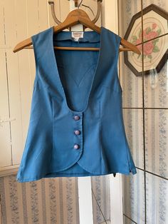 This pretty vest is made from permanent press fabric in a beautiful shade of medium blue. It has the original covered buttons and western style detailing on the back yoke. Very fitted. The measurements, taken with the vest lying flat, are: shoulder to shoulder, 12 1/2 inches; armpit to armpit, 15 inches; length, 22 inches in front and 22 inches in back; bottom edge, 17 inches. In very good condition. Fitted Classic Cotton Denim Vest, Fitted Blue Cotton Denim Vest, Classic Fitted Cotton Denim Vest, Fitted Cotton Denim Vest With Buttons, Classic Fitted Denim Vest For Spring, Fitted Cotton Vest With Button Closure, Fitted Denim Vest With Button Closure For Work, Blue Denim Vest For Work With Buttons, Fitted Sleeveless Denim Vest With Button Closure