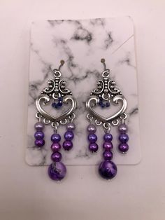 2 Inch drop-earrings made with hypoallergenic materials.   Beads are attached to a silver plated heart charm consisting of a purple marble bead and three smaller glass beads in three different shades of purple/lavender in the center.  Left/Right sides contain the same smaller glass beads in the three different shades of purple/lavender as the center.  The middle of the heart charm is accented with four tiny glass beads as an accent.  These beads are a darker shade of purple. Includes soft ear backings. Materials are environmental, lead free and non-toxic. Light weight, smooth surface, and a touch of sheen to add elegance. The earrings are attached to a marble cardboard earring holder and are shipped in a sealed plastic cover, and placed inside a mesh drawstring "thank you" pouch as shown. Purple Teardrop Chandelier Earrings As Gift, Purple Drop Chandelier Earrings As Gift, Handmade Purple Sterling Silver Chandelier Earrings, Purple Dangle Heart Earrings For Pierced Ears, Purple Teardrop Metal Earrings, Purple Heart-shaped Beaded Jewelry, Purple Metal Dangle Earrings, Purple Dangling Beads Chandelier Earrings, Nickel-free Purple Beaded Earrings