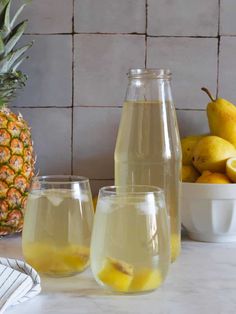 two glasses filled with lemonade next to a pitcher and bowl of fruit on a counter