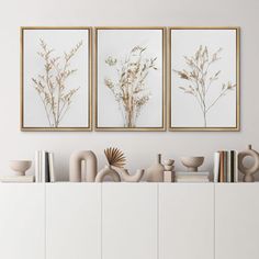 three framed pictures on the wall above a white cabinet with books and vases in it