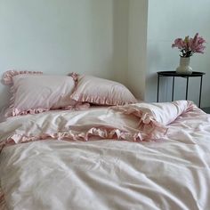 a white bed with pink ruffles and pillows in a room next to a table