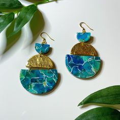 two pairs of blue and gold earrings on top of a white table next to green leaves