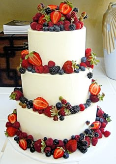 a three tiered white cake with berries and strawberries on the top is sitting on a table