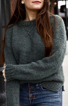 a woman with long brown hair wearing a green sweater and glasses, standing in front of a building