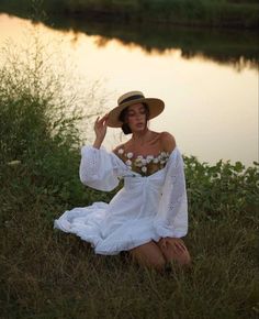 a woman in a white dress and hat sitting on the grass near water at sunset