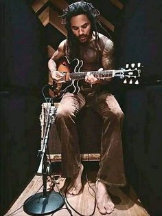 a man with dreadlocks sitting on a chair playing an acoustic guitar in front of a microphone