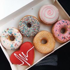 six donuts in a white box with sprinkles and heart shaped frosting