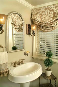 a white sink sitting under a mirror next to a window with roman shades on it