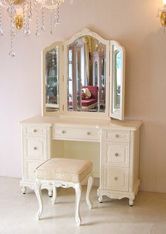 a white dressing table with a mirror and stool in front of it, next to a chandelier