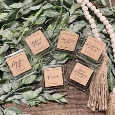 four small square candles sitting on top of a wooden table next to green leaves and a tassel