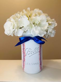 white flowers in a baseball jar with blue ribbon and name on the side, sitting on a table